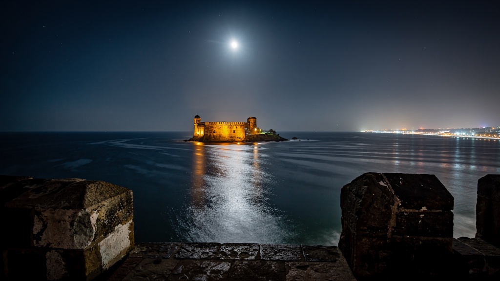 Panikotha glowing at night seen across the dark water from the main fort